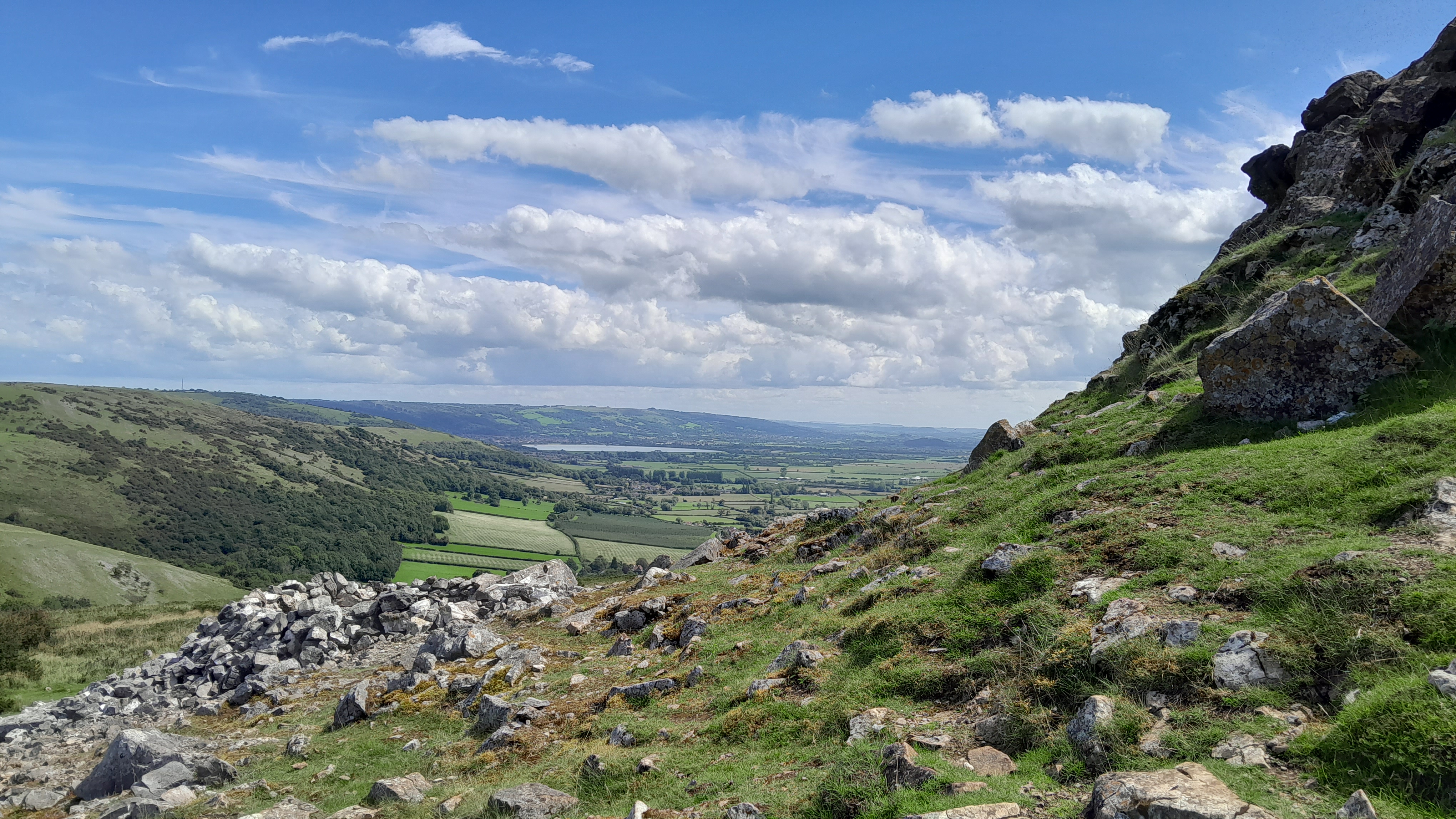 View from near the top of a craggy rock looking out across green fields and moorland to a circular reservoir